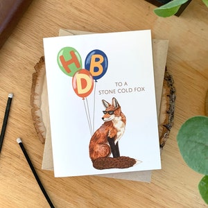 May include: A birthday card with a fox wearing sunglasses and holding balloons that spell out "HBD". The text on the card reads "To a Stone Cold Fox".