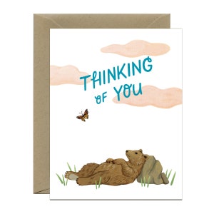 May include: A greeting card with a brown bear lying on its back with its paws in the air. The bear is resting on a rock with green grass around it. A butterfly is flying in the upper left corner of the card. The text "Thinking of you" is written in blue on a white background with pink clouds.