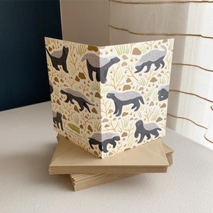 May include: A card with a pattern of honey badgers on a white background. The badgers are black and gray with white stripes. The background is a light yellow with brown branches and small yellow flowers.