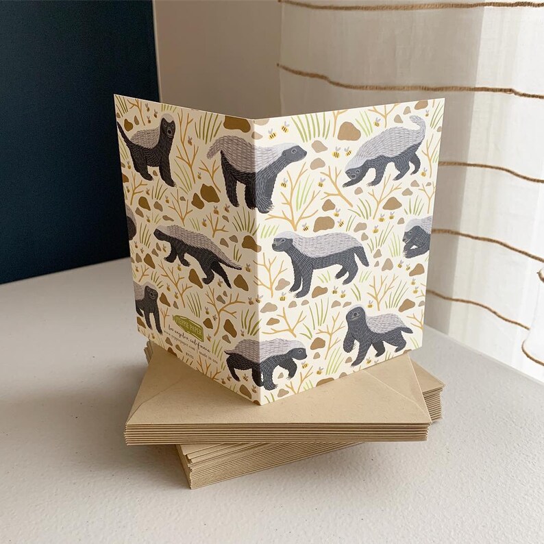 May include: A card with a pattern of honey badgers on a white background. The badgers are black and gray with white stripes. The background is a light yellow with brown branches and small yellow flowers.