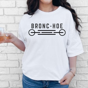 Puede incluir: Una camiseta blanca de manga corta con el texto negro BRONC-HOE impreso en el pecho. Debajo del texto hay un diseño gráfico negro que consiste en una línea horizontal con círculos en los extremos y tres líneas paralelas en el centro.