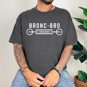 Peut inclure: Un t-shirt gris anthracite à manches courtes avec le texte blanc BRONC-BRO centré sur la poitrine, placé au-dessus d'un motif graphique horizontal composé de deux cercles reliés par trois lignes parallèles.