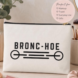 Puede incluir: Un estuche rectangular de lona color blanco roto con cremallera, que presenta el texto negro BRONC-HOE centrado sobre un gráfico de una línea horizontal que conecta dos círculos. El estuche tiene una cremallera negra y un tirador dorado.
