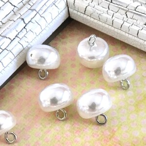 May include: A collection of 15 white pearl buttons with silver loops. The buttons are arranged in a frame on a pink and white patterned surface. A penny is included for size reference.