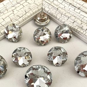 May include: A collection of clear, faceted crystal buttons arranged on a white surface within a distressed white frame. The buttons vary in size and reflect light, creating a sparkling effect. A silver button is also visible.