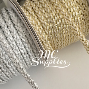 May include: Two spools of braided trim, one gold and one silver. The trim is a woven, rope-like material with a subtle sheen.