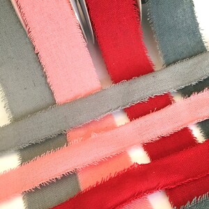May include: Three spools of fabric ribbon in gray, pink, and red. The ribbon is frayed on the edges and has a rustic look.