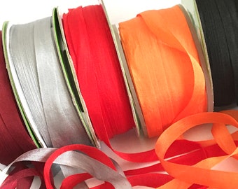 May include: Assortment of colorful silk ribbon spools in burgundy, silver, red, orange, and black. The ribbon is unraveled and laying on a white surface.