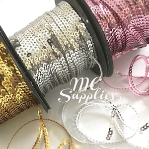 May include: Three spools of sequined trim in gold, silver, and pink. The spools are on a white background. The trim is perfect for adding a touch of sparkle to any project.