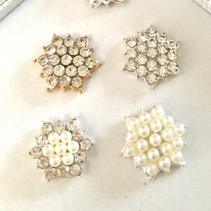 May include: Five decorative snowflake embellishments in a variety of styles. Two are silver with clear rhinestones, one is gold with clear rhinestones, and two are silver with white pearl accents.