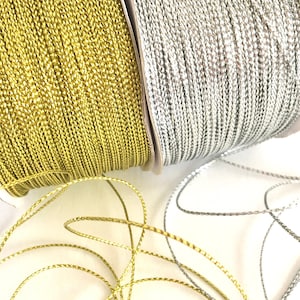 May include: Two spools of metallic thread, one gold and one silver. The thread is twisted and has a subtle shimmer. The spools are on a white background.