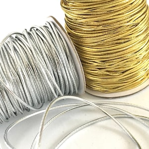 May include: Two spools of metallic elastic cord, one silver and one gold. The silver cord is unwound and lying on a white surface.