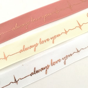 May include: Three satin ribbons with a gold foil heartbeat design and the text "always love you". The ribbons are pink, cream, and white.