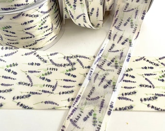 May include: A set of five spools of lavender-patterned ribbon. The ribbon is white with a purple lavender print. The ribbon is on a white surface.