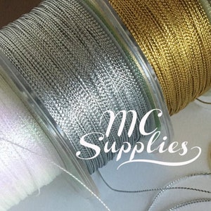 May include: Three spools of metallic thread, one gold, one silver, and one white. The spools are on a white surface with the thread unraveled in circles.