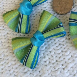 May include: Five green and blue striped fabric bows with a turquoise center. The bows are arranged on a white knitted surface.