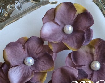 May include: Close-up of several purple fabric flowers with yellow edges and a pearl center. The flowers are arranged on a clear, decorative glass plate. The text "MC Supplies" is visible in the image.