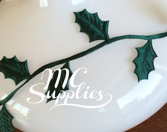 May include: Green holly leaf trim with a white vase. The trim is made of fabric and has a satin finish.