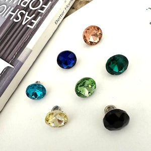 May include: Assortment of seven decorative glass buttons in various colours, including blue, green, yellow, black, and peach. The buttons are round and have a faceted design.