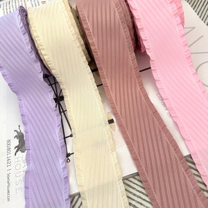 May include: Four rolls of ribbon with a ruffled edge and a diagonal stripe pattern. The ribbons are in shades of purple, cream, pink, and brown.