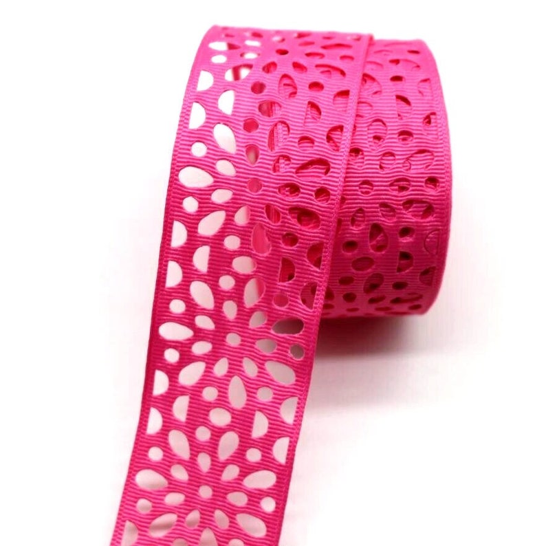 May include: Pink satin ribbon with a floral cut-out design. The ribbon is rolled up and the cut-out design is visible on the edge.