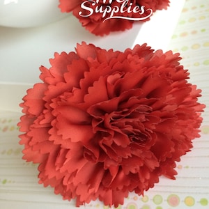 May include: A close-up of a red fabric carnation flower. The flower is made of soft, ruffled fabric and has a realistic appearance.