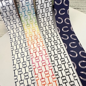 May include: Four rolls of white ribbon with different patterns. The first roll has a black and white pattern of horseshoes. The second roll has a rainbow pattern of horseshoes. The third roll has a black and white pattern of horseshoes. The fourth roll has a navy blue background with pink horseshoes.