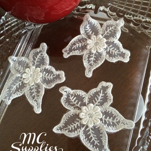 May include: Three white lace flower appliques with a delicate floral design. The appliques are on a clear glass surface with a brown background. The text "MC Supplies" is visible in the bottom left corner of the image.