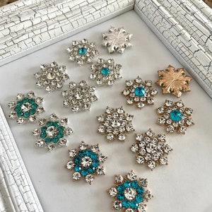 May include: Assortment of snowflake-shaped embellishments with clear and turquoise rhinestones. The embellishments are arranged in a white frame.
