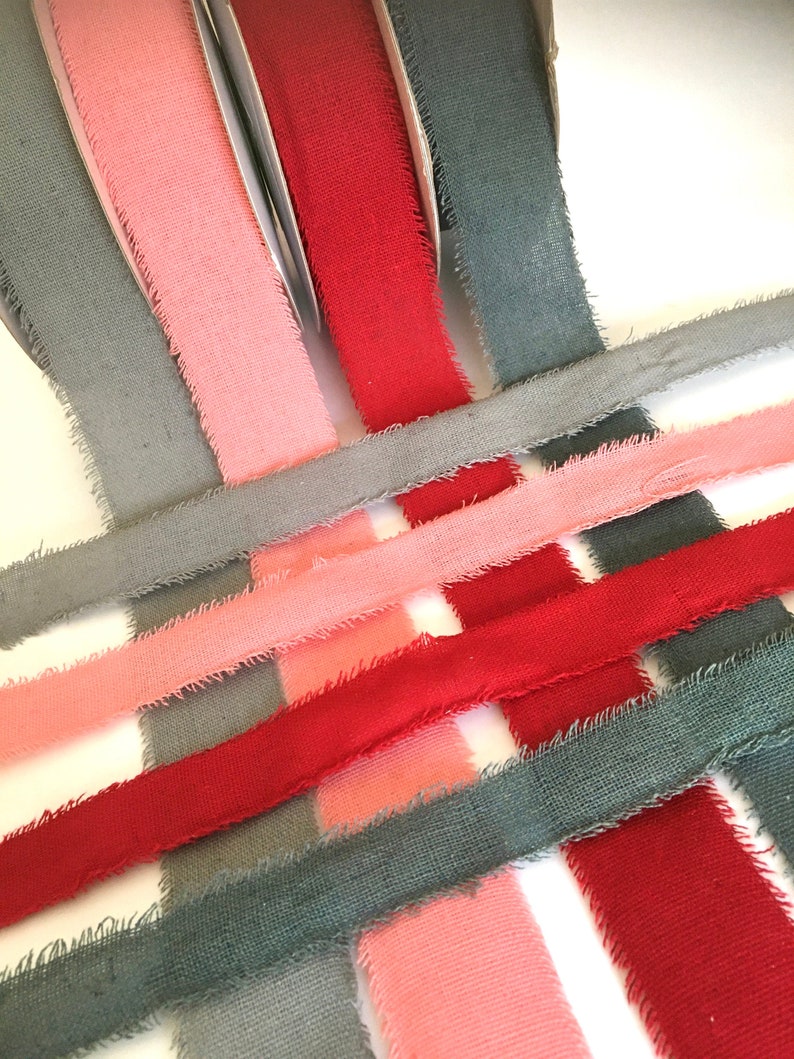 May include: Three spools of fabric ribbon in gray, pink, and red. The ribbon is frayed on the edges and has a rustic look.