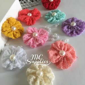 May include: Assortment of nine small, handmade fabric flowers with pearl centers. The flowers are in various colors, including red, pink, yellow, white, light blue, and purple. The flowers are arranged on a white surface. MC Supplies is written in cursive on the surface.