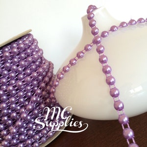 May include: A spool of lavender pearl trim with a single strand of the trim draped over a white vase. The trim is made of small, round pearls that are strung together on a thin wire. The trim is perfect for adding a touch of elegance to any project.