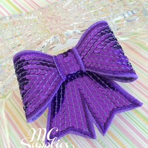 May include: A purple sequined bow with a satin finish. The bow is on a striped background.