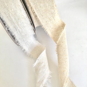 May include: Two rolls of natural linen ribbon with frayed edges. One roll is white and the other is a light beige.