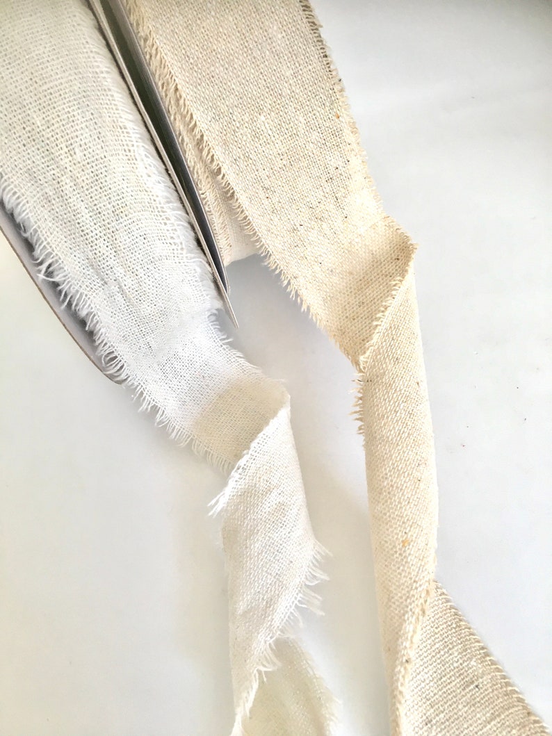 May include: Two rolls of natural linen ribbon with frayed edges. One roll is white and the other is a light beige.