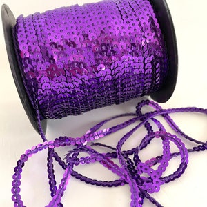 May include: A spool of purple sequin trim. The trim is made of small, round sequins that are sewn together on a thin thread. The sequins are shiny and have a metallic finish.