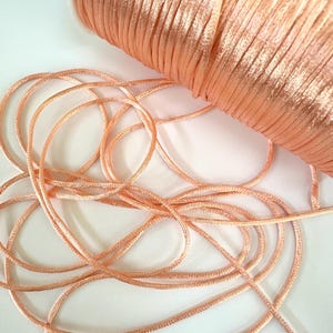 May include: A spool of peach-colored satin cord with loose strands. The cord is tightly wound on the spool, and the loose strands are scattered on a white surface. The satin cord has a slight sheen, reflecting light.