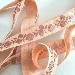May include: Peach-colored elastic ribbon with a rose pattern in a metallic copper color. The ribbon is folded and twisted, showing the pattern from different angles.