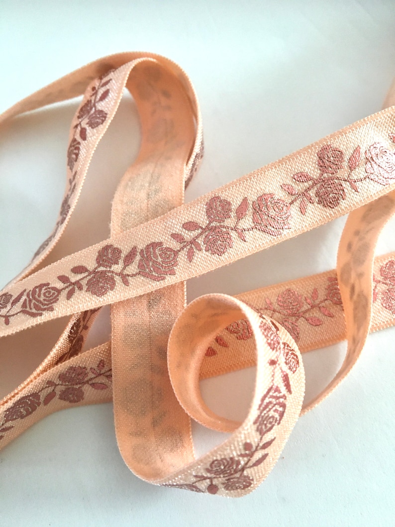 May include: Peach-colored elastic ribbon with a rose pattern in a metallic copper color. The ribbon is folded and twisted, showing the pattern from different angles.