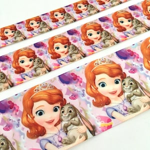 May include: Pink satin ribbon featuring a repeating pattern of a princess with brown hair and a crown holding a brown bunny. The princess is wearing a purple dress with a light blue sash. The background is a light pink with purple flowers and white swirls.