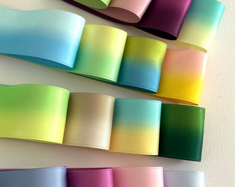 May include: A collection of wide satin ribbons arranged in horizontal rows, featuring various gradient color combinations including pink to green, blue to yellow, lime to gold, and purple to lavender, with a smooth, reflective, and lustrous finish.
