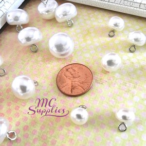 May include: A collection of white pearl buttons with silver loops. The buttons are arranged on a pink and white polka dot background. A penny is included for scale.