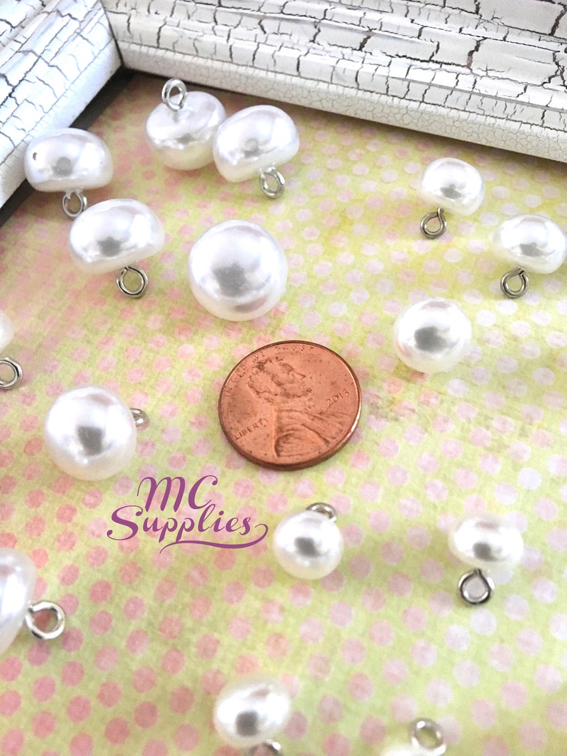 May include: A collection of white pearl buttons with silver loops. The buttons are arranged on a pink and white polka dot background. A penny is included for scale.