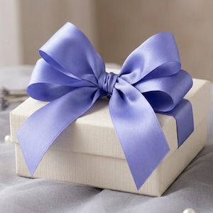 May include: A square, off-white gift box featuring a smooth, satin-finish lavender ribbon tied into a large, multi-looped bow on the lid. The ribbon wraps around the sides of the box, creating a clean, structured presentation for a small item.