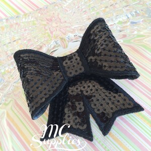 May include: A black sequined bow with a classic shape. The bow is made of shiny, black sequins and has a stitched border. The text "MC Supplies" is visible at the bottom of the image. The bow is likely used for crafting or embellishment.