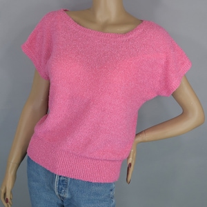 May include: A bright pink, short-sleeved sweater with a wide, boat neckline. The sweater has a textured, fuzzy appearance and a ribbed hem. The garment is displayed on a mannequin, paired with blue jeans.