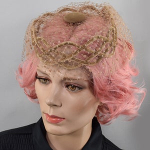 May include: A vintage hat with a delicate net veil and a decorative crown. The hat features a beige net and a small, round, beige accent on top. The mannequin has pink hair and is wearing a black top.