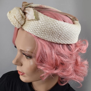 May include: A vintage cream-colored hat with a woven texture and a decorative bow. The hat features a band that wraps around the head and is accented with a light-colored fabric and a small floral detail. The hat is a classic accessory.