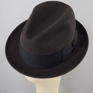 May include: A dark brown fedora hat with a black band. The hat has a classic, slightly curved brim and a pinched crown. The hat is displayed on a mannequin head against a plain background.