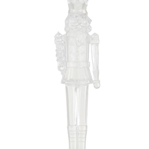 May include: A clear, transparent plastic nutcracker figurine. The figurine is detailed with a crown, uniform, and boots. The nutcracker stands on a small, round base. The overall design is a classic holiday decoration.